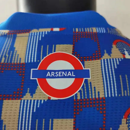 Maglia arsenal joint edition 2022/23 blu con logo circolare rosso e bianco e motivo edifici beige su sfondo blu reale